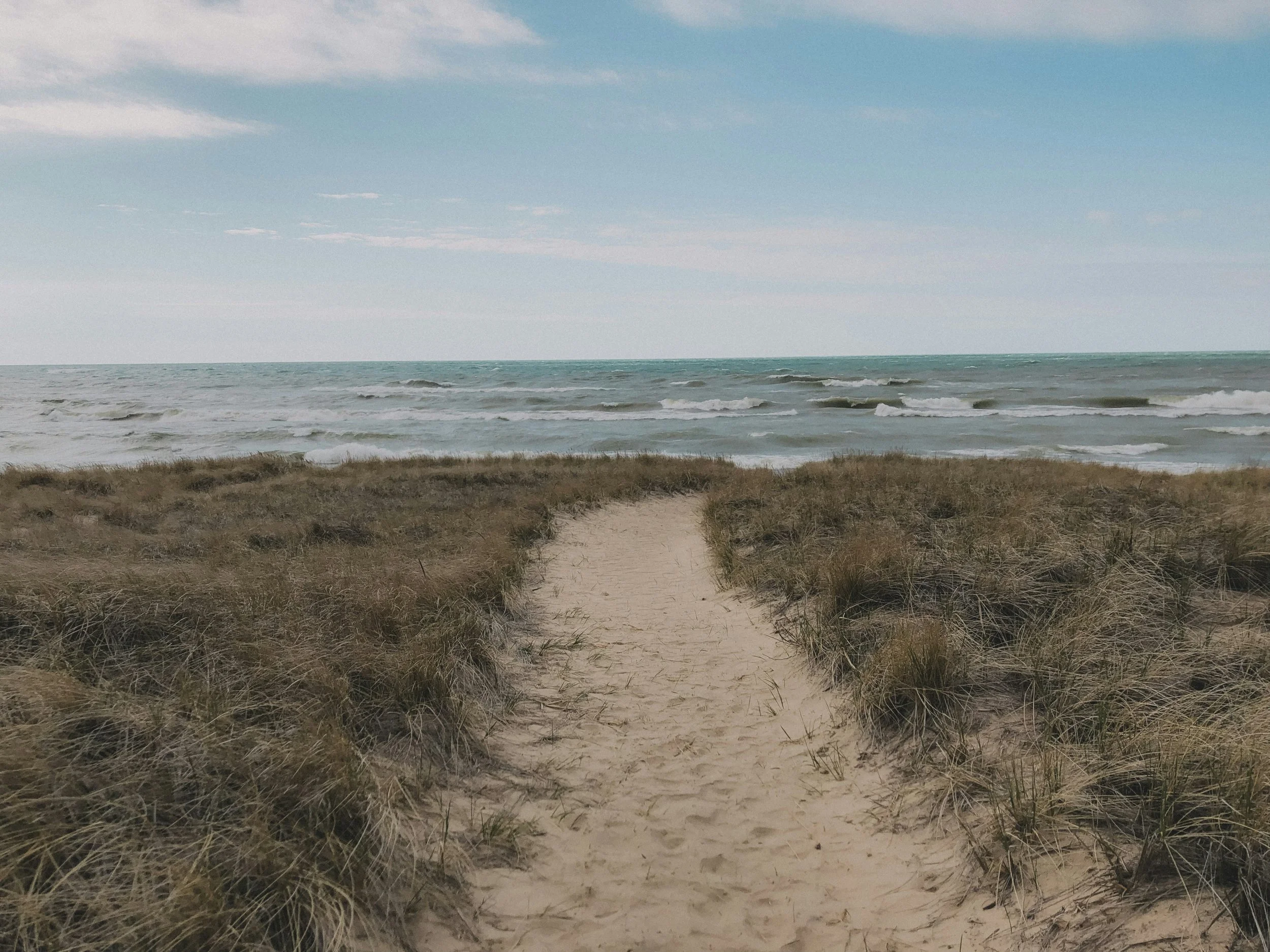 A sandy path leading to the ocean, flanked by grassy dunes under a partly cloudy sky.