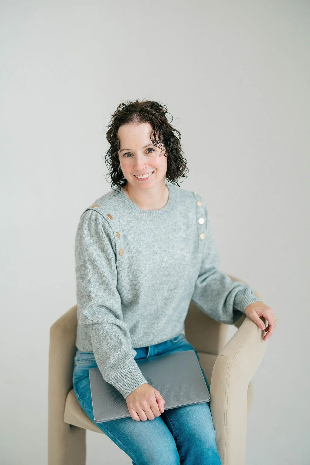 Jileah, Online Business Manager: a woman with dark curly hair, wearing a gray sweater with gold buttons on the shoulders, sitting on a beige chair, holding a closed laptop on her lap, smiling at the camera against a plain light gray background.