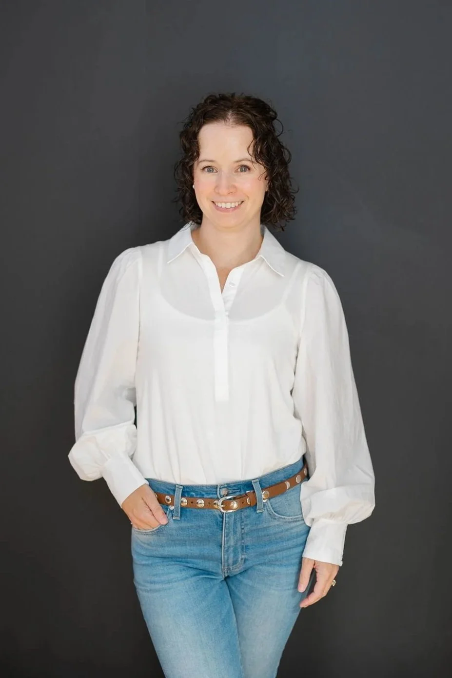 Jileah, Online Business Manager: a woman with short curly brown hair wearing a white blouse and blue jeans standing against a dark background.