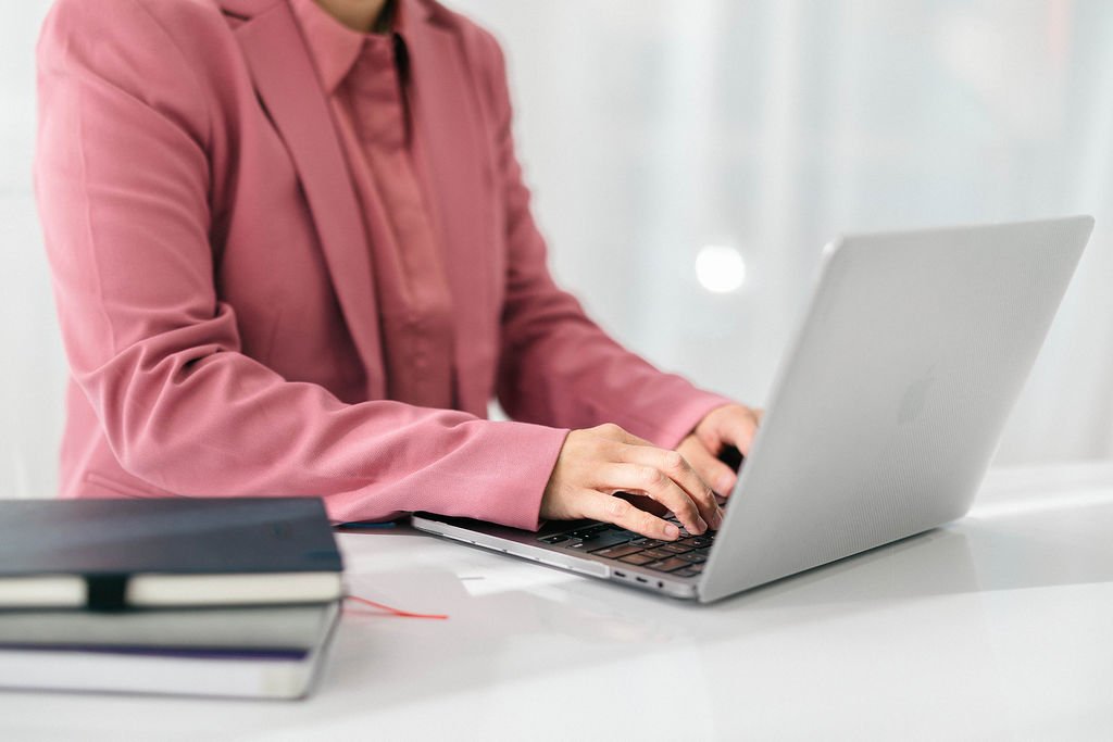 Person in pink blazer working on a silver laptop at a white desk with a closed black notebook nearby.