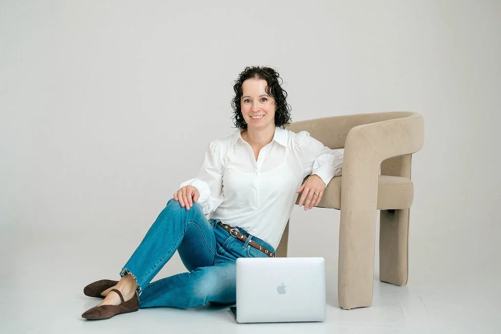 Woman with curly black hair, white blouse, blue jeans, and brown shoes sitting on the floor next to a beige armchair and a silver laptop, in front of a plain background.