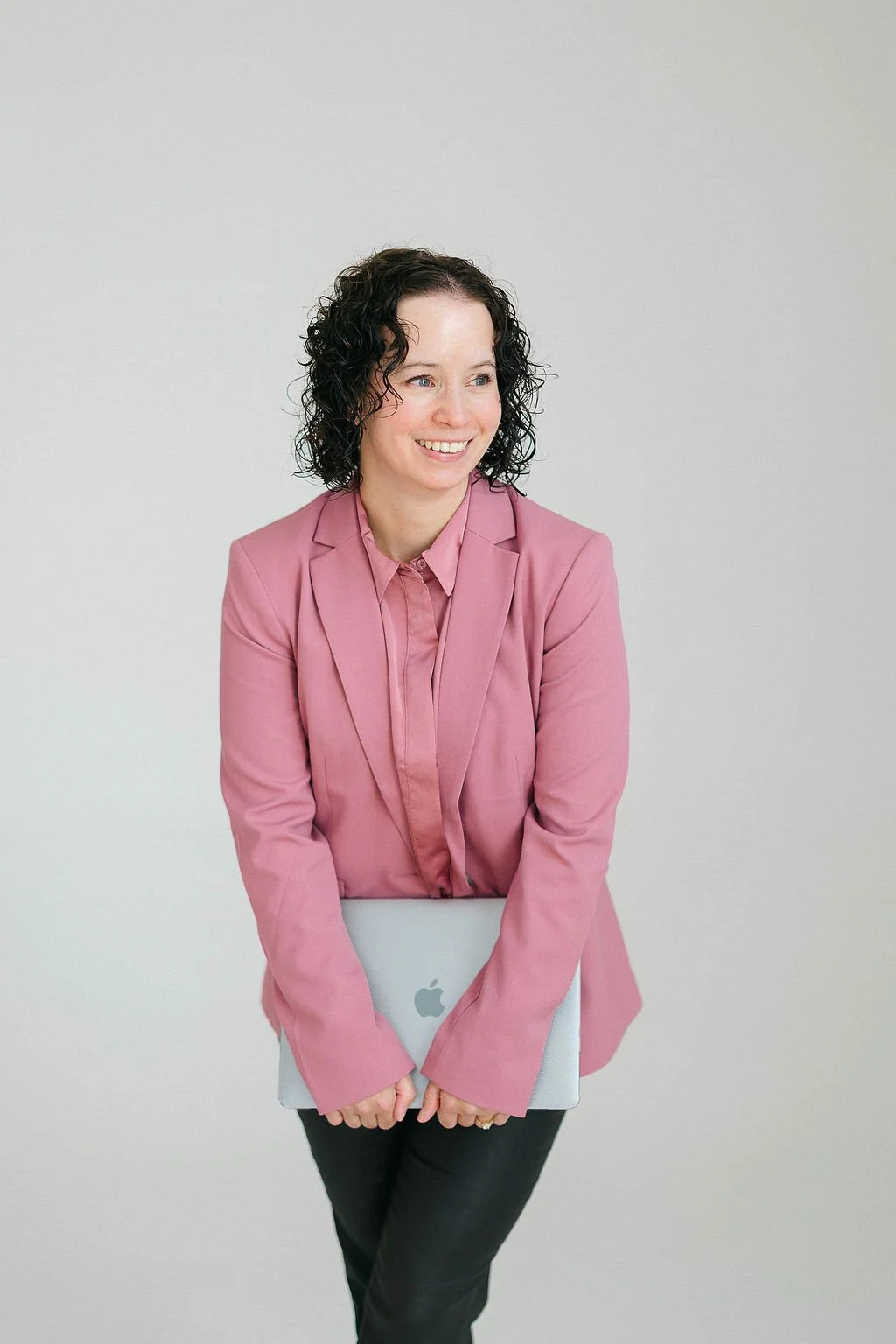 Jileah, Online Business Manager: a woman with curly dark hair wearing a pink blazer and blouse, holding a closed silver MacBook, standing against a plain gray wall, smiling.