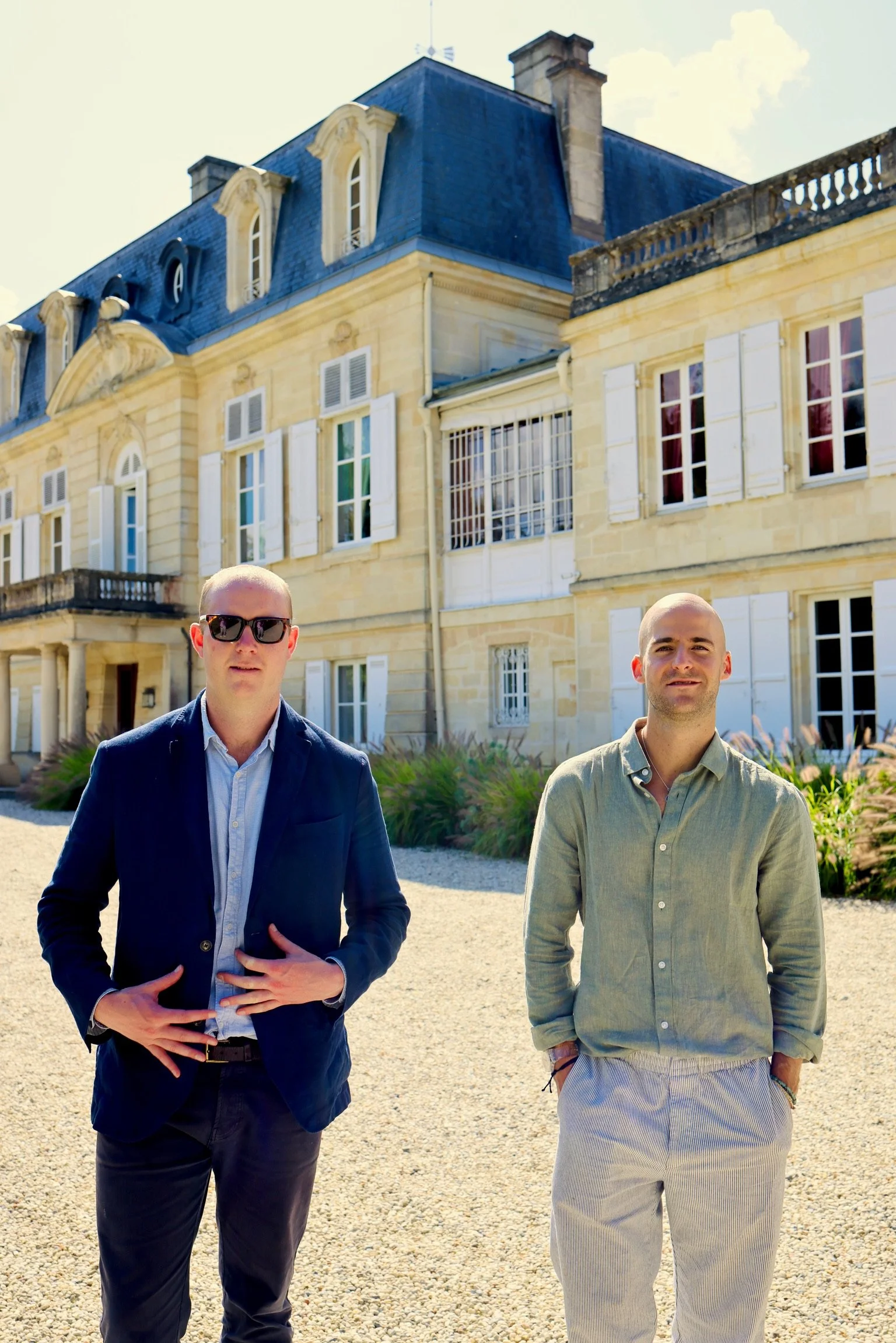 Two men standing outdoors in front of a large, elegant, historic building with beige stone walls and white window shutters. One man is wearing a dark blazer and sunglasses, and the other is wearing a light-colored shirt and pants. The sky is clear and sunny.