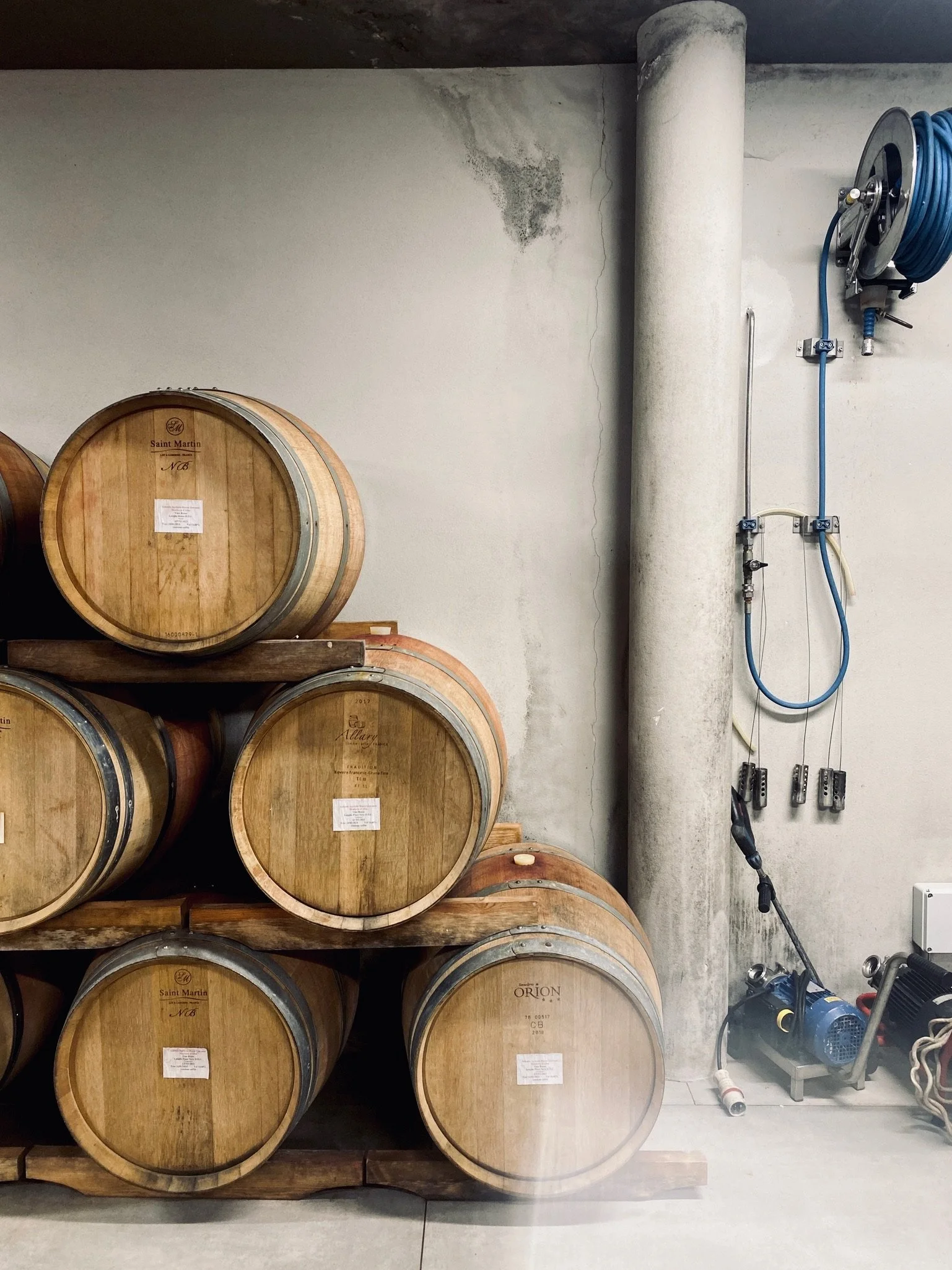 Group of wine barrels stacked on a wooden rack in a cellar with a plumbing hose reel and equipment on the wall.