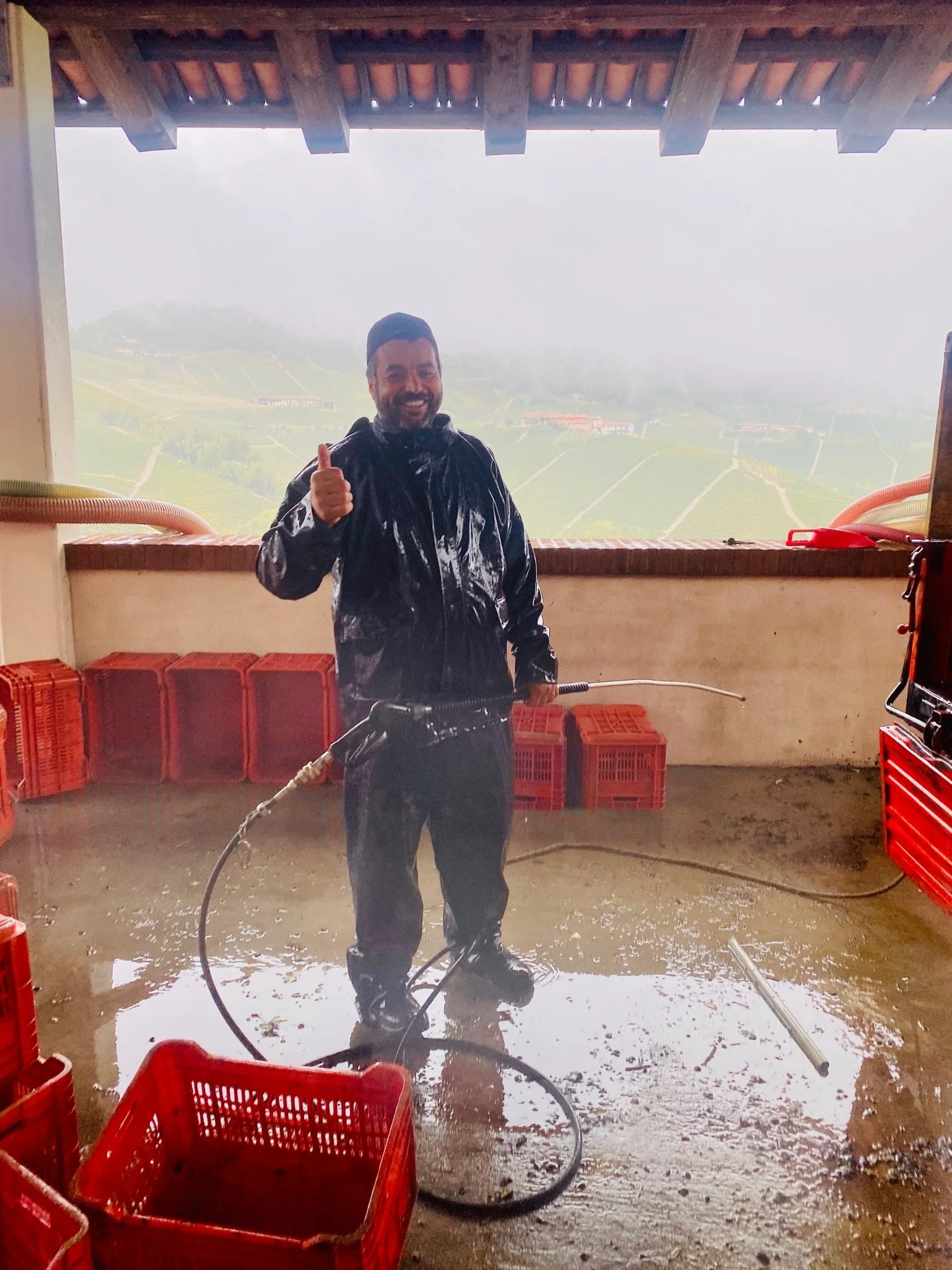 A man in a black raincoat holding a pressure washer in a room with red plastic crates and a misty landscape in the background, giving a thumbs-up.