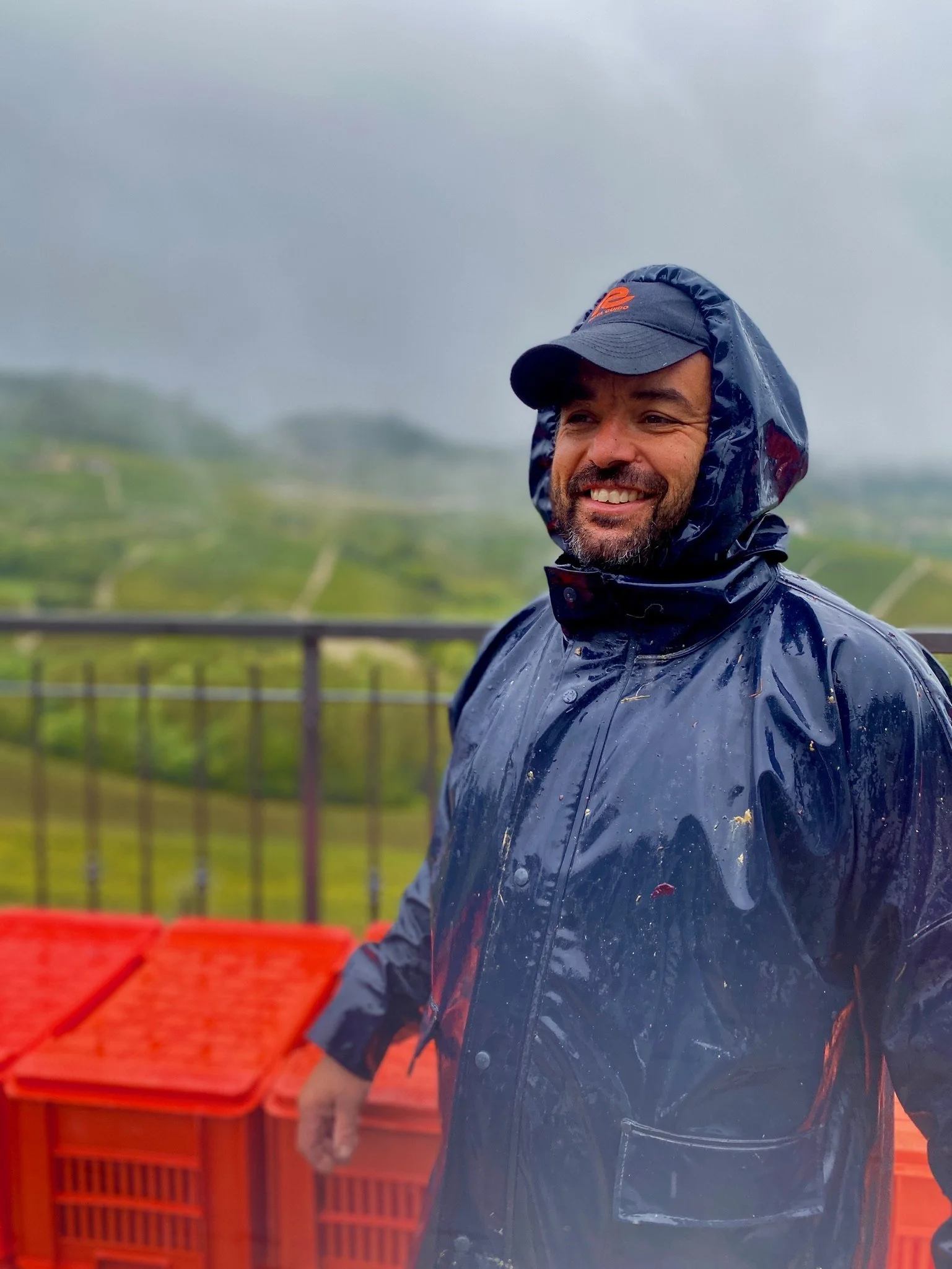 A man smiling outdoors on a rainy day, wearing a dark blue rain jacket with a hood and a black cap, with a blurred green landscape in the background.