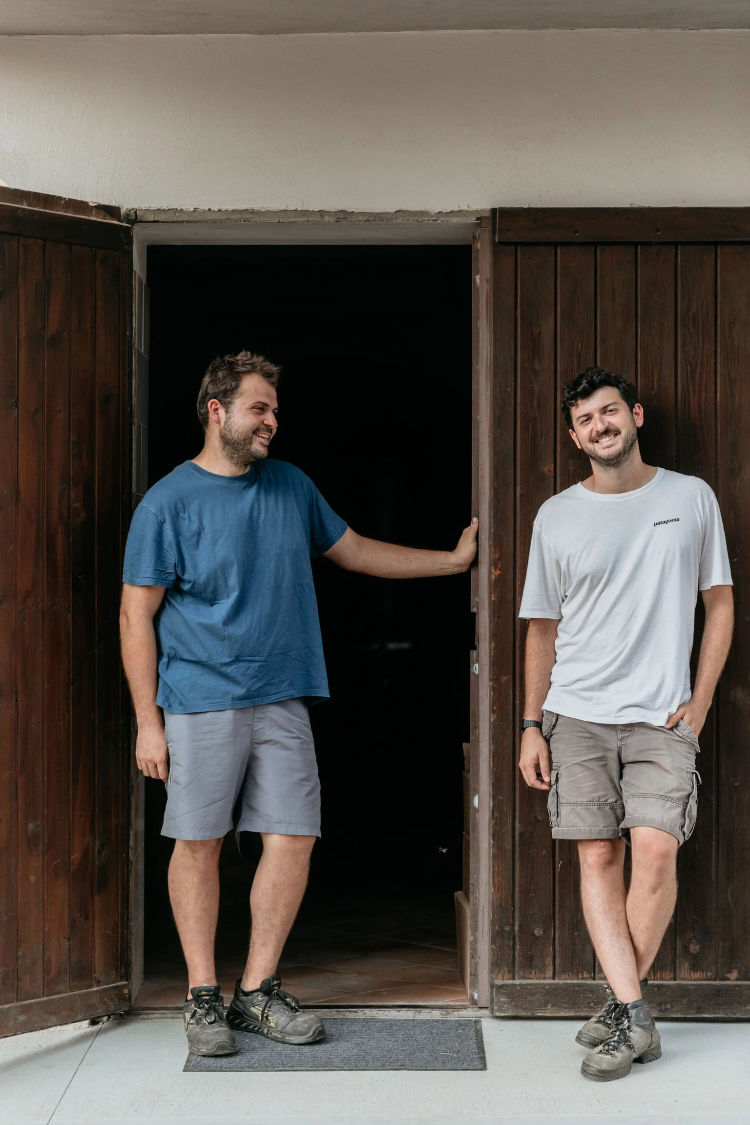 Two young men casually standing outside a wooden door, one leaning against the doorframe and the other standing with hands in pockets, smiling.