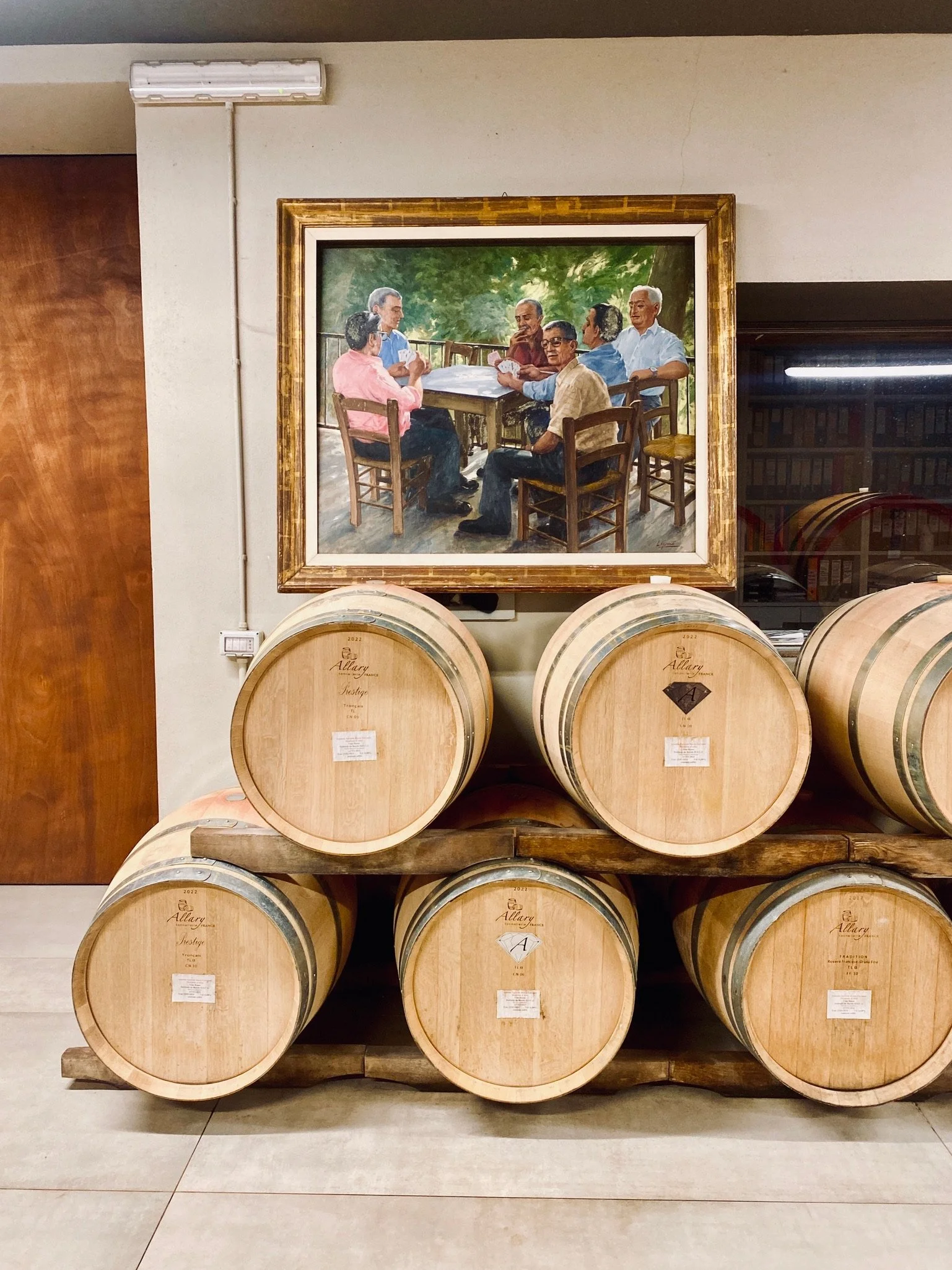 A group of six men sitting around an outdoor table in a painting, with some playing cards, surrounded by green trees.