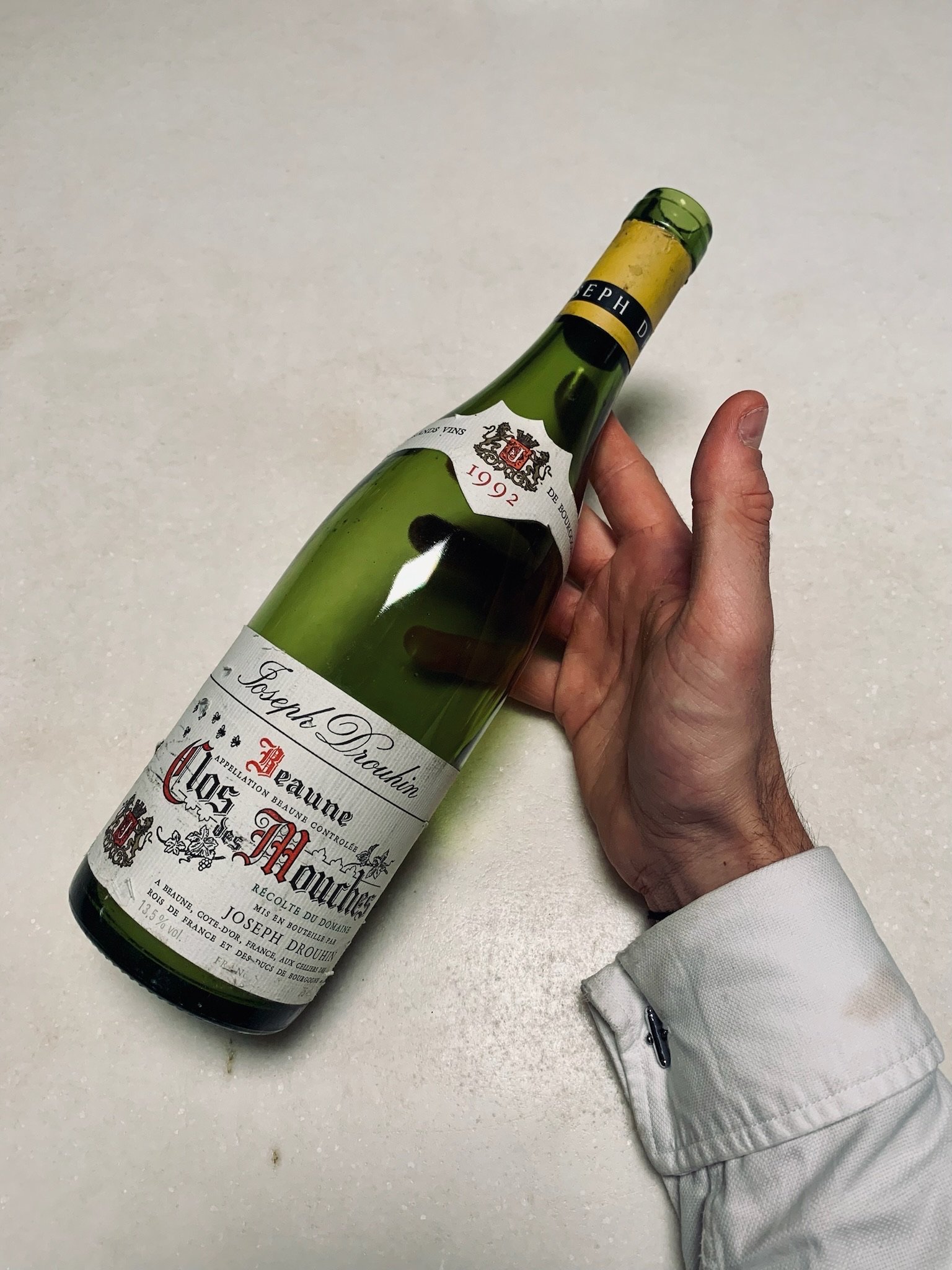 A hand holding a green wine bottle labeled 'Joseph Drouhin Clos des Mouches' on a white surface.