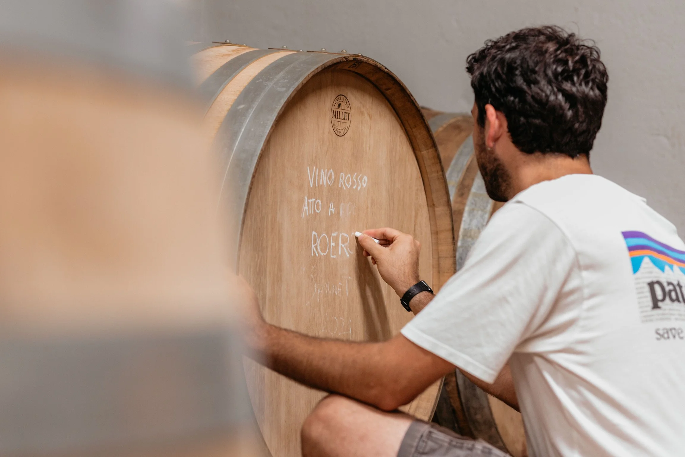 A man writing on a wooden wine barrel with white chalk in a winery.