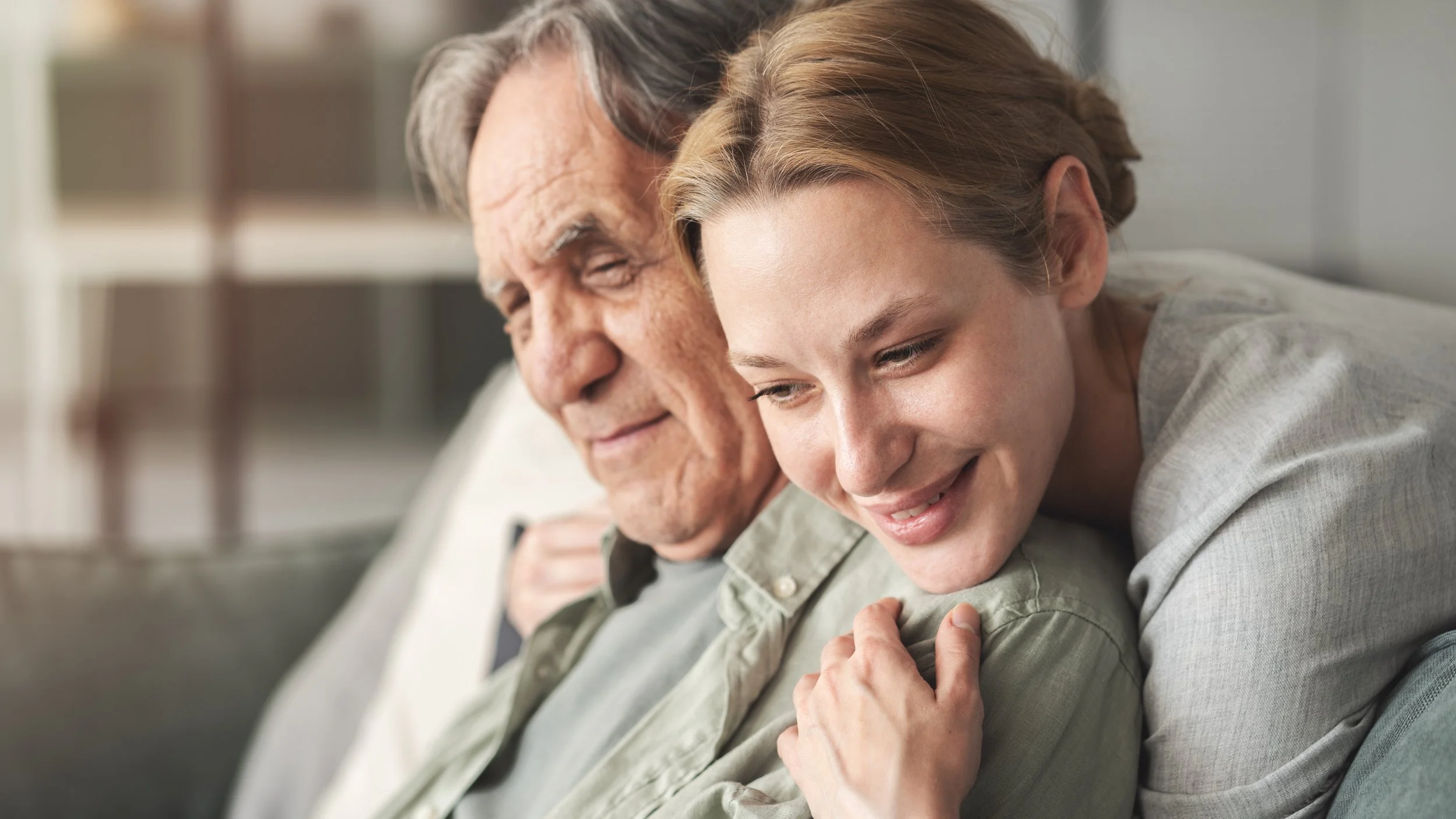 An elderly man and a young woman sitting close together on a couch, smiling and sharing a warm moment.