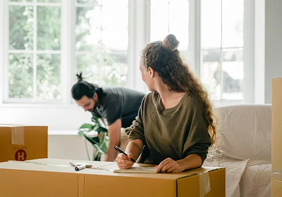A woman sitting at a table in a bright room, looking away while a man in the background arranges a box.