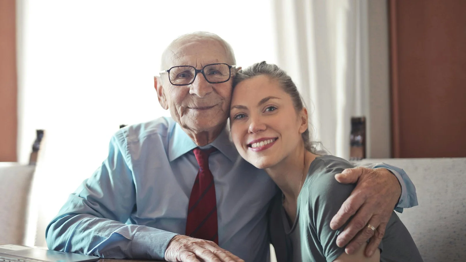 An elderly man wearing glasses and a light blue shirt, and a young woman with long hair, smiling and hugging, sitting together indoors in a well-lit room.