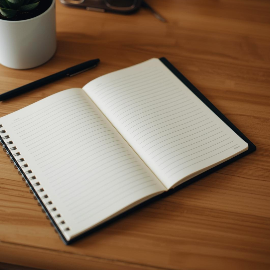 Open blank notebook with lined pages on wooden desk, next to a black pen and a potted plant in a white pot.