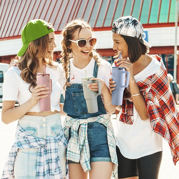 Three young women smiling and enjoying drinks at an amusement park or fair during daytime, with a red and white striped structure in the background.