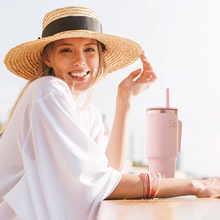 Young woman smiling, wearing a large straw hat with black band, sitting outdoors at a table with a pink reusable tumbler and straw.