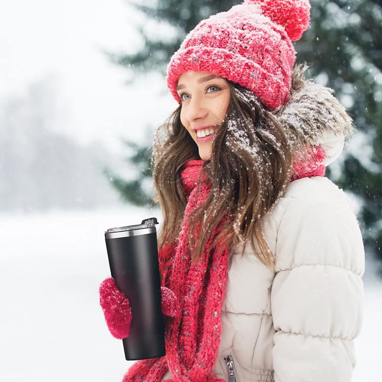 Smiling woman in winter clothing holding a travel mug outdoors in a snowy landscape