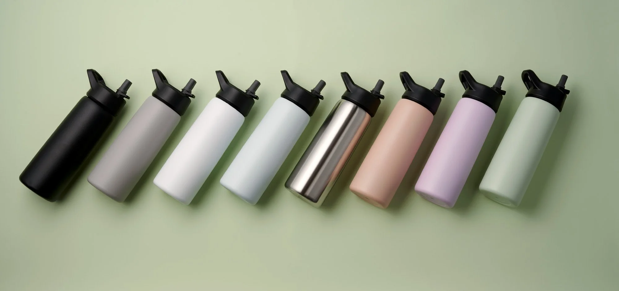 A row of eight reusable water bottles in various colors including black, gray, white, metallic, beige, purple, and mint green, lined up on a light green background.