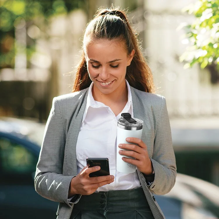 A young woman in business attire smiling while looking at her smartphone outdoors, holding a reusable coffee cup.