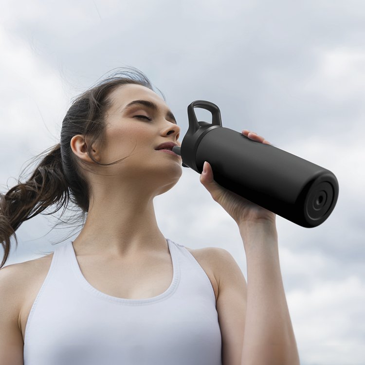 A young woman with long hair, wearing a white tank top, drinking water from a black water bottle outdoors on a cloudy day.
