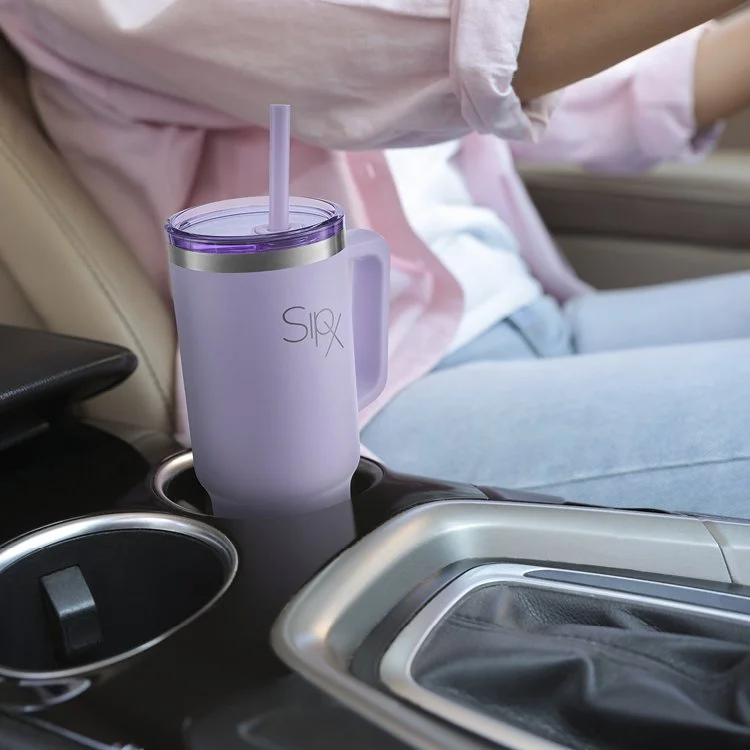 A purple travel mug with a straw in a car's cup holder. There is a person in a pink shirt and blue jeans sitting in the passenger seat.
