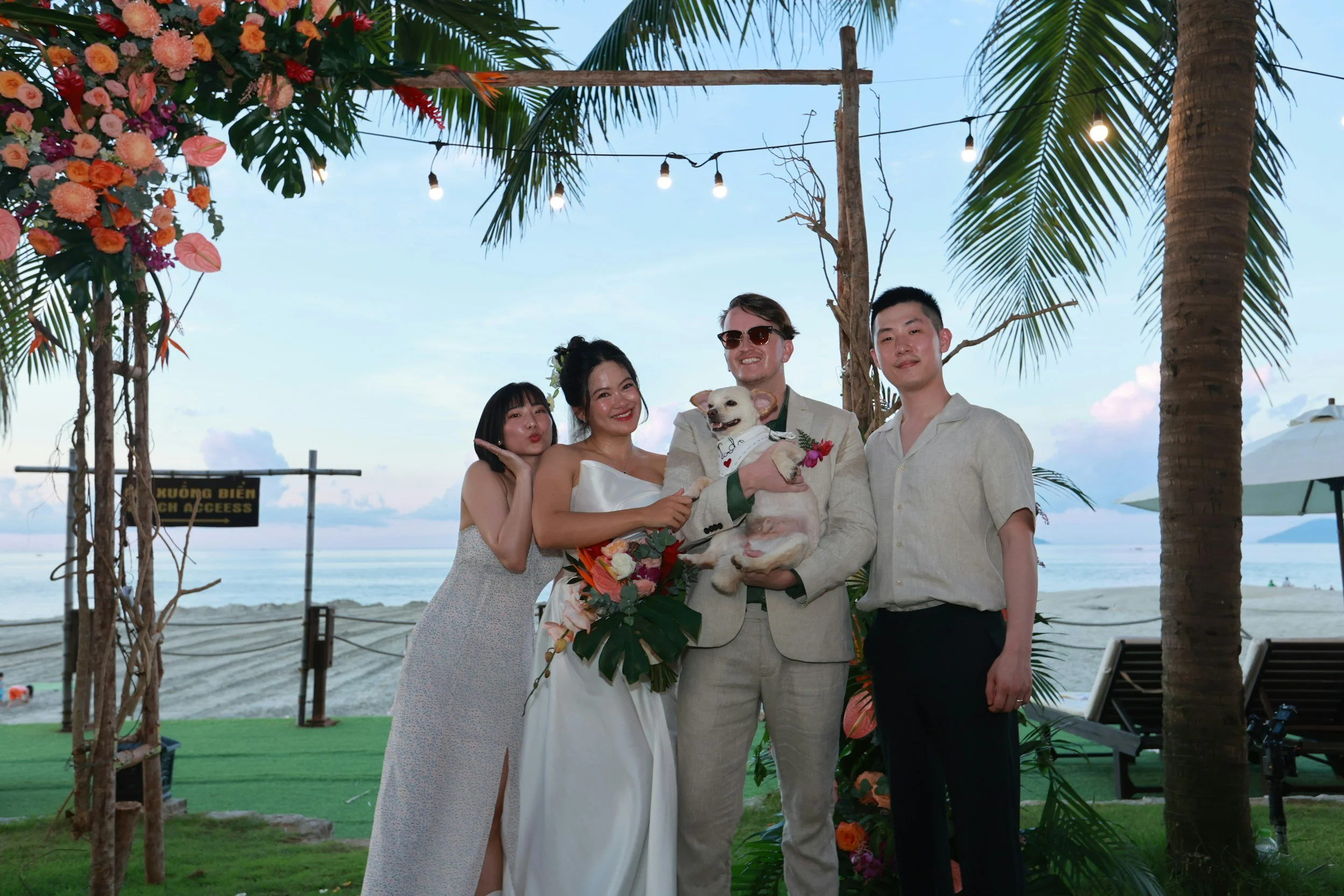 A group of five friends at a beachside wedding celebration, with palm trees, flowers, and string lights overhead, posing happily with a dog in the groom's arms.