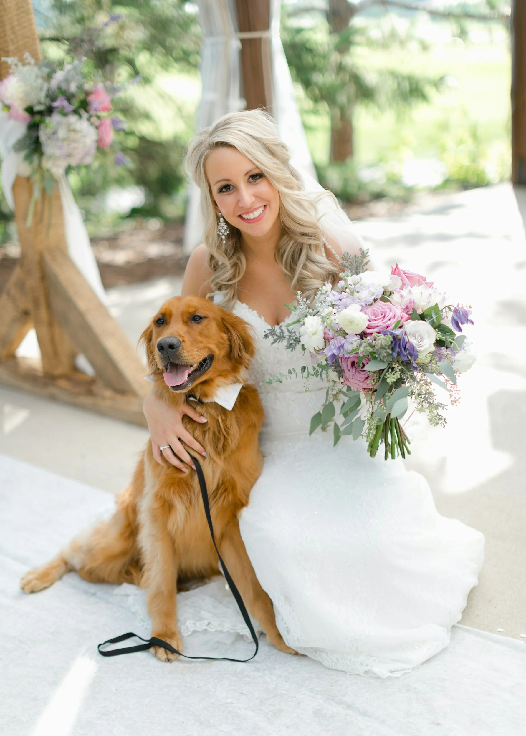 A bride in a white wedding dress, holding a large bouquet of pink, white, and purple flowers, sitting indoors near a wooden arch decorated with flowers, with a golden retriever dog sitting beside her.