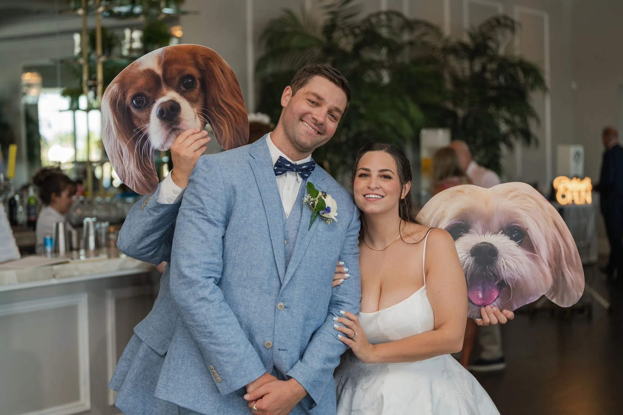 A newlywed couple at their wedding reception, smiling and posing for a photo with two large cutouts of dogs, one of a Cavalier King Charles Spaniel and the other a Shih Tzu, in the background.