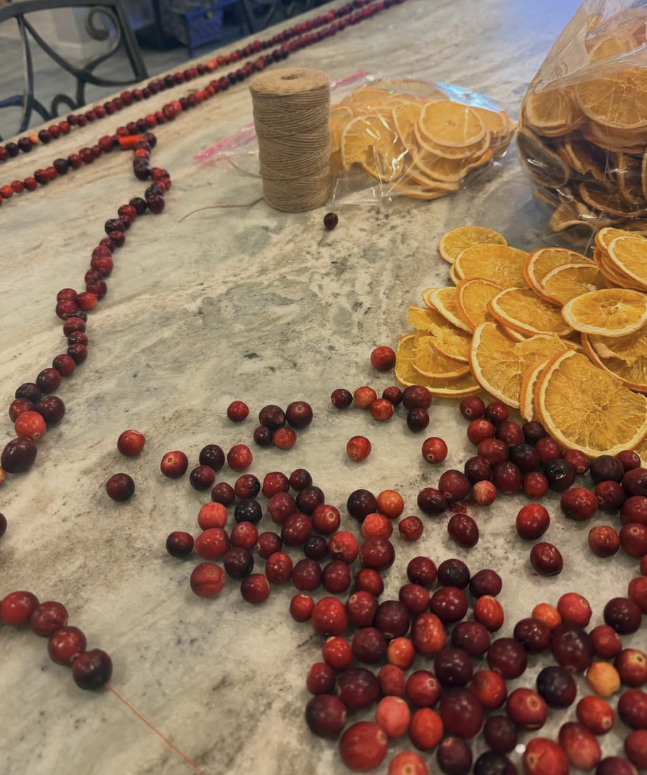 Unfinished cranberry bead jewelry with dried orange slices, a spool of string, and plastic bags of dried oranges on a marble countertop.
