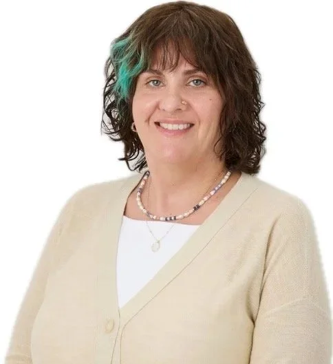 A woman with shoulder-length curly brown hair with a turquoise streak, smiling, wearing a beige sweater, a white top, a pearl necklace, and a necklace with a circular pendant, against a white background.