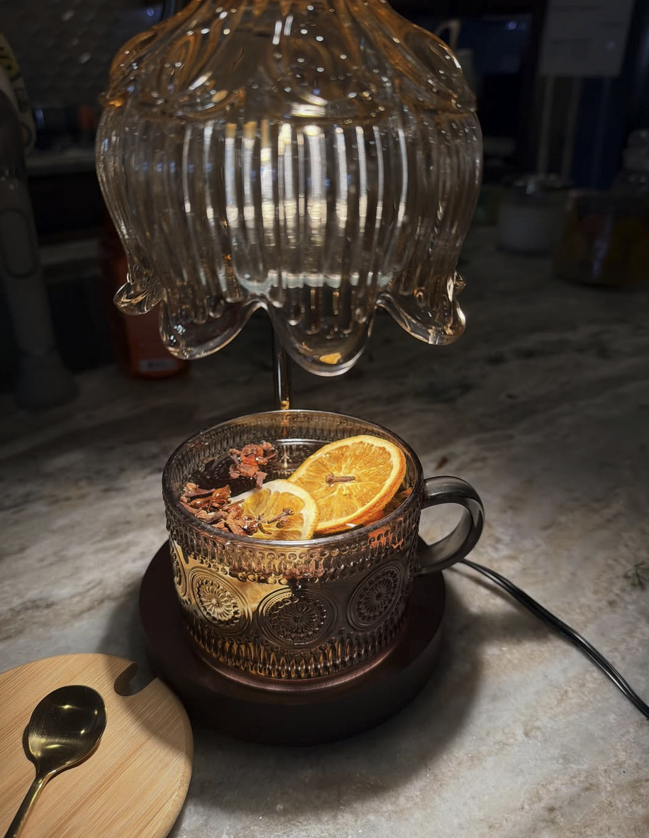 A glass teacup with lemon slices and herbs inside, placed on a wooden tray, with a decorative glass lamp overhead, on a marble countertop.