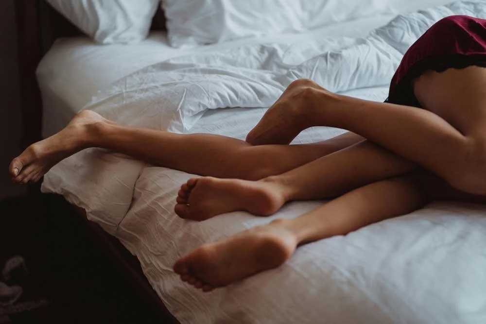 Close-up of two people lying on a bed, intertwined with their legs and feet visible, with white bedding and a dark headboard.