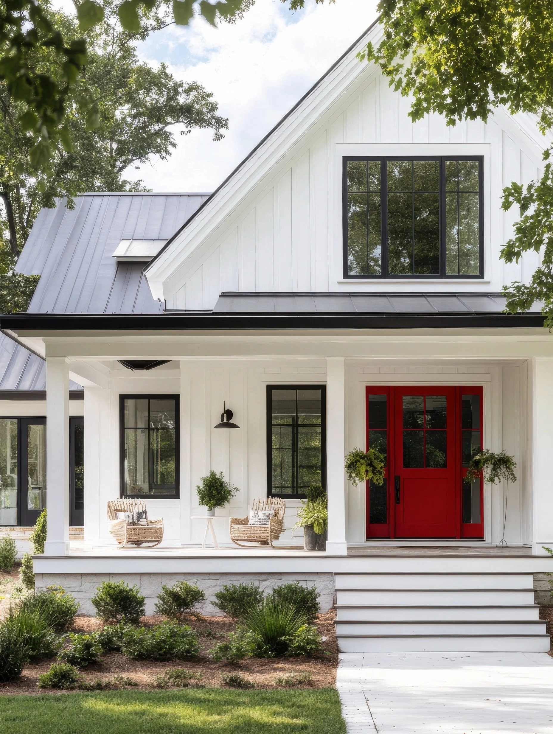 A modern white house with a red front door, black window frames, and a porch with outdoor furniture and plants, set amidst greenery.
