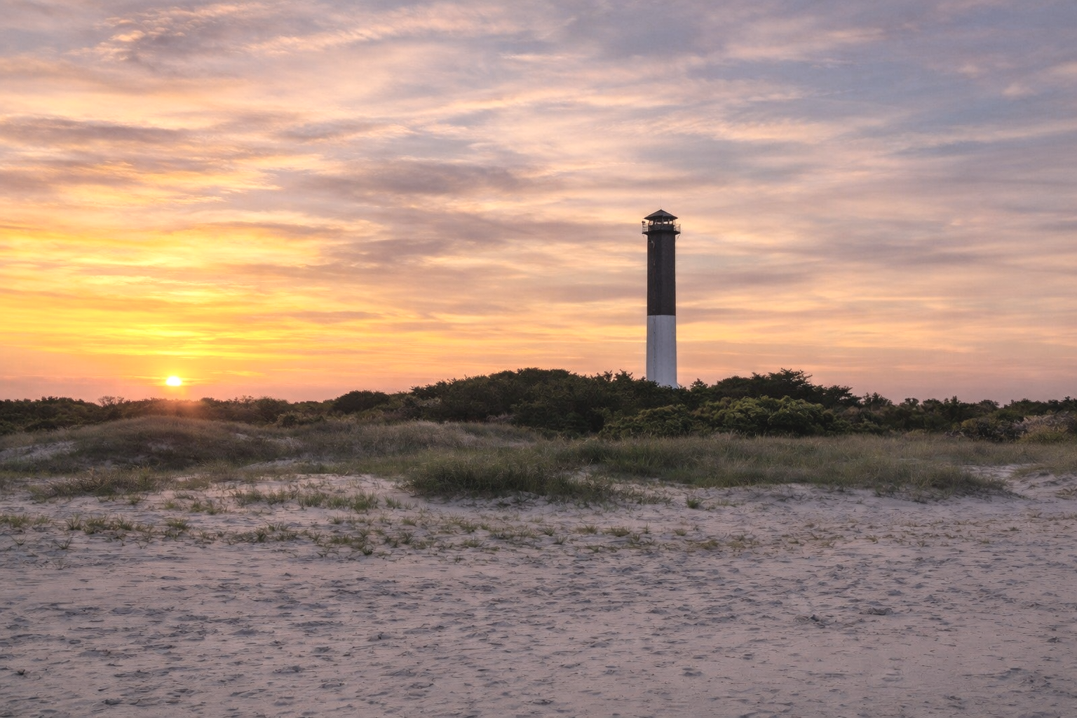 A lighthouse stands tall on a small hill covered with shrubbery, with the sun setting behind it, casting an orange and pink glow across the sky, and sandy dunes in the foreground.