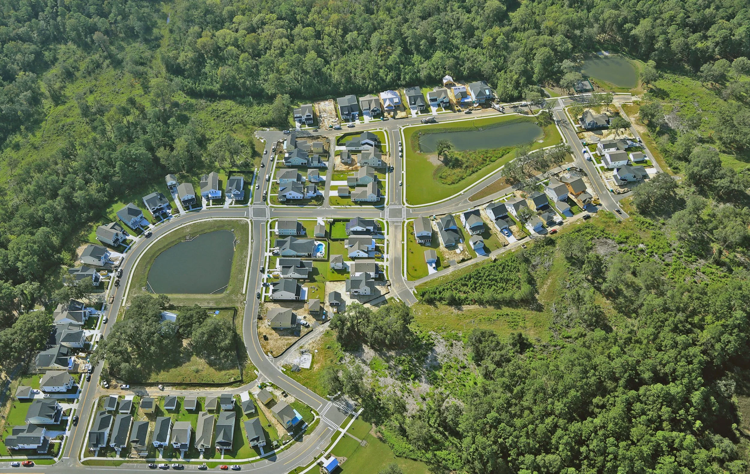 Aerial view of a suburban neighborhood with houses, two ponds, streets, and surrounded by dense green forest.