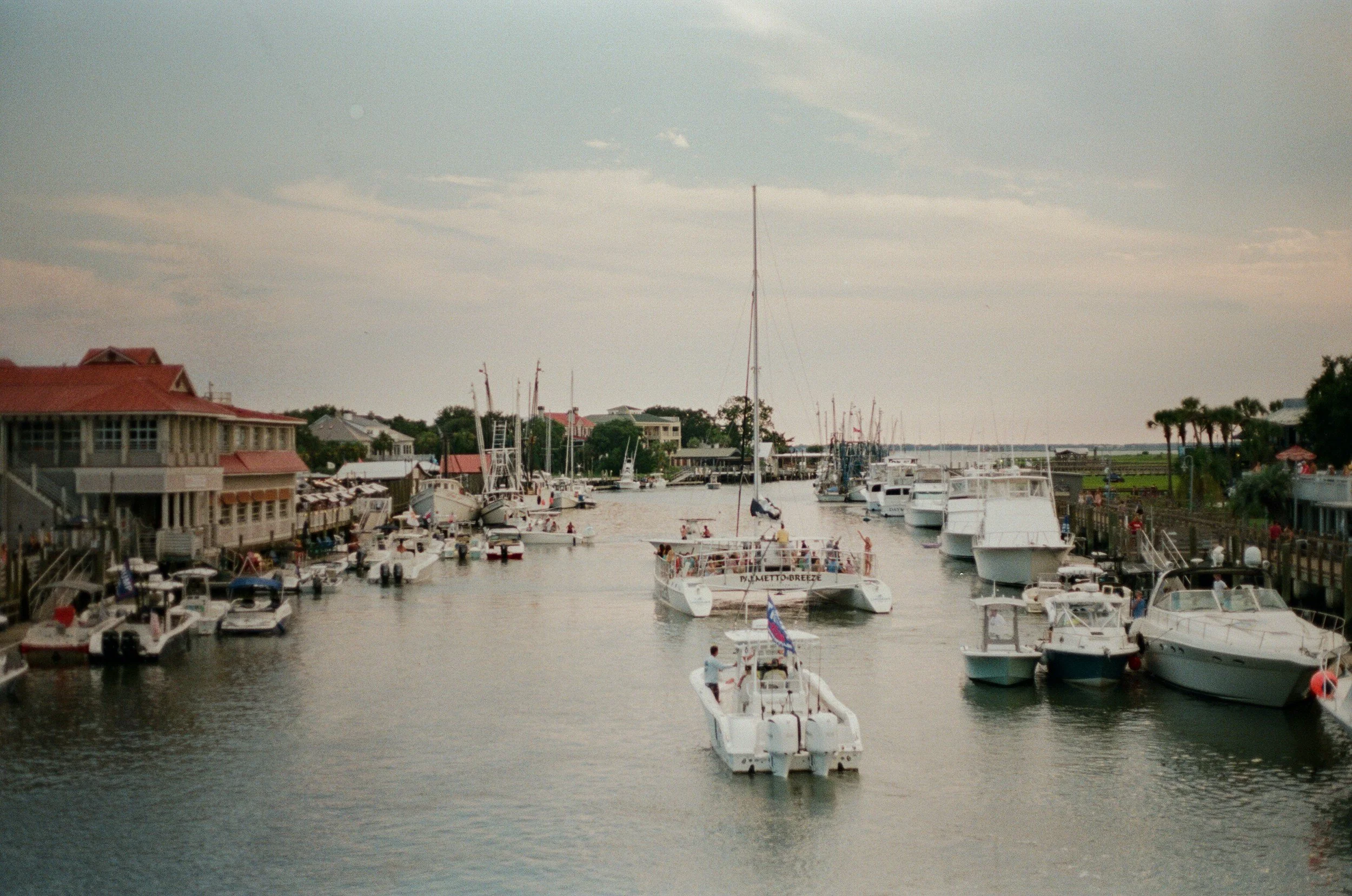 A marina with various boats and yachts docked on both sides, and a boat sailing in the center of the waterway. There are buildings with red roofs on the left and some palm trees on the right, under a partly cloudy sky.