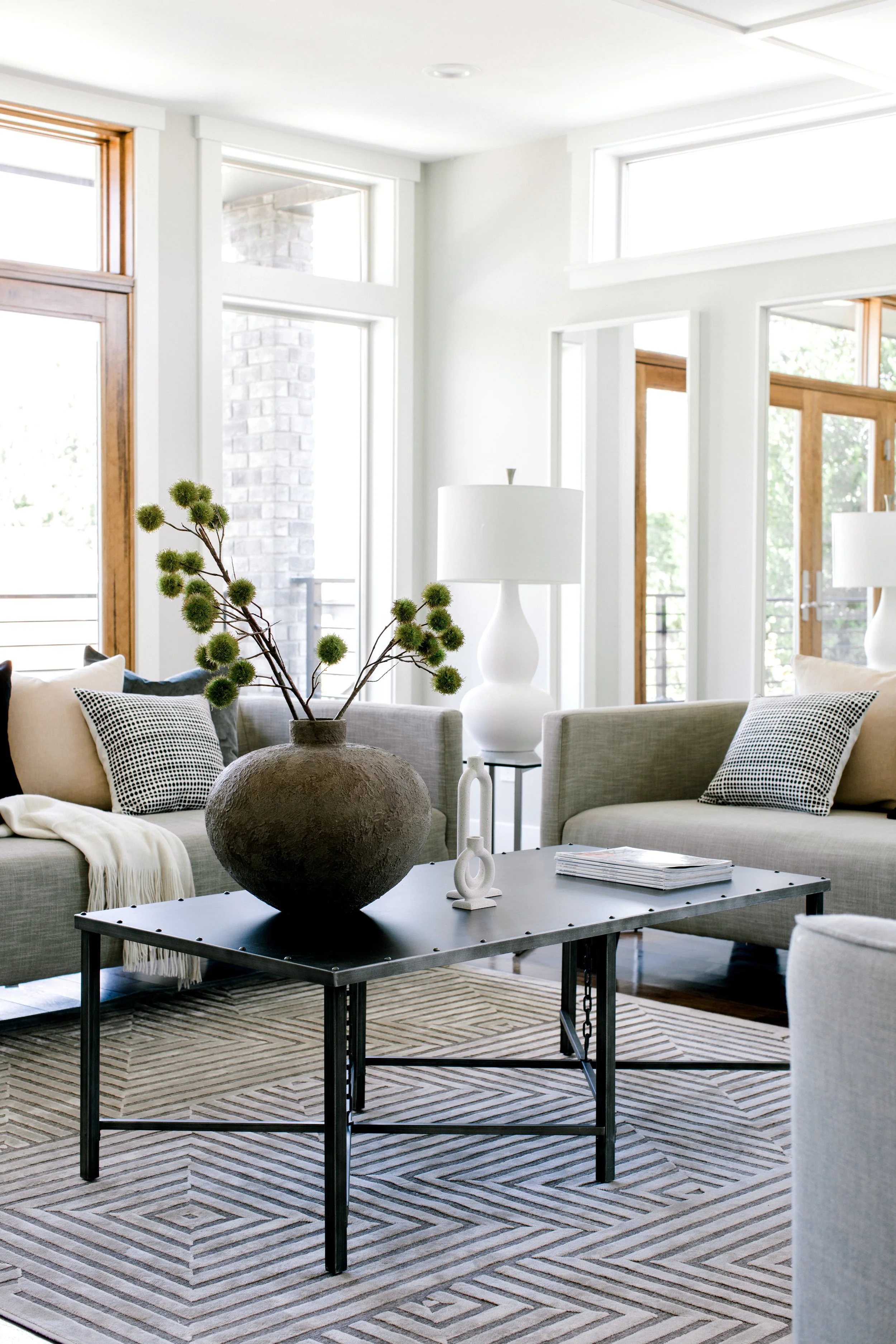 Modern living room with large windows, gray sofas with black and white pillows, a black coffee table with a large vase and a white sculpture, and beige patterned rug.