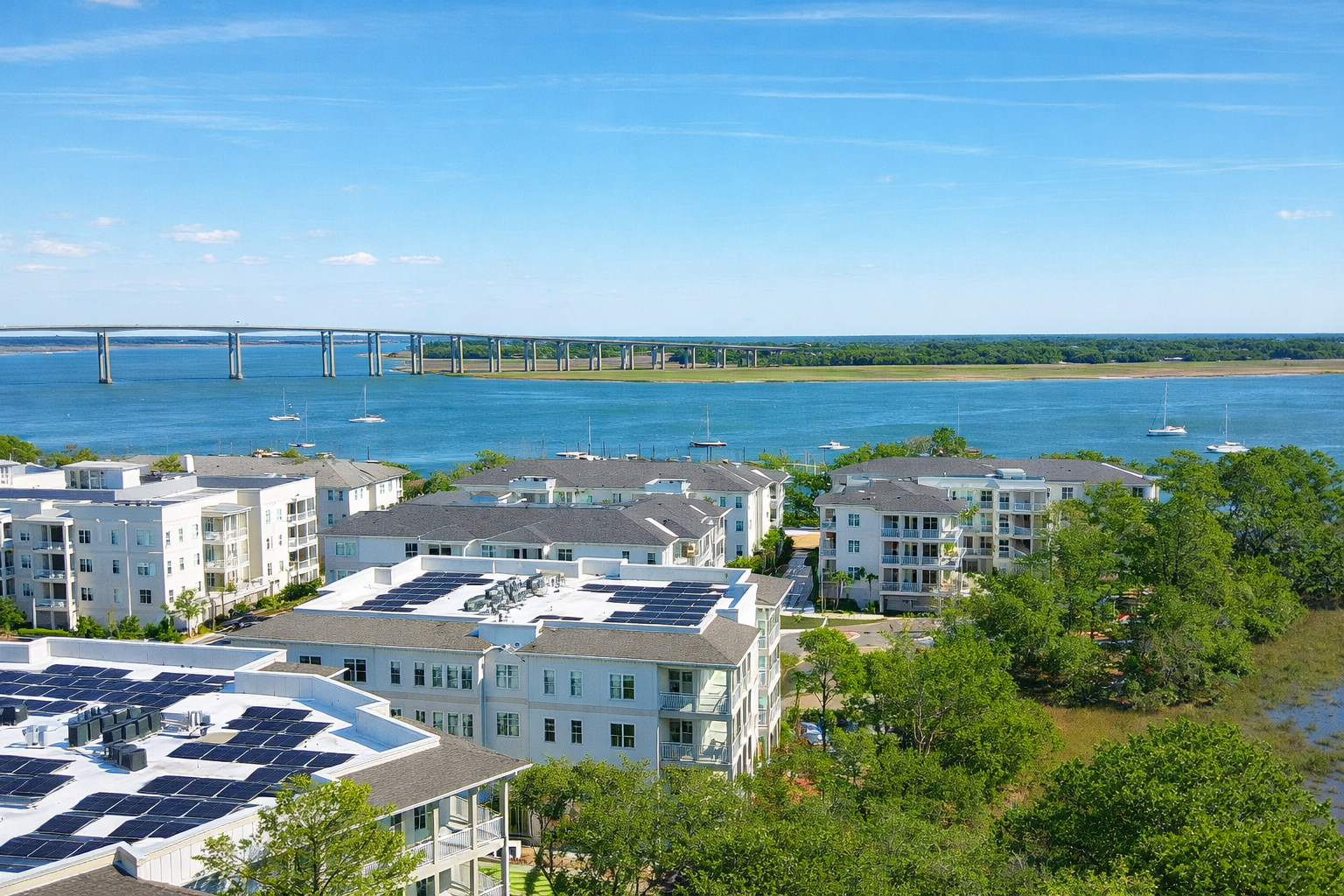 View of white apartment buildings with solar panels on roofs, green trees, and a large body of water with sailboats, and a bridge in the background under a partly cloudy blue sky.