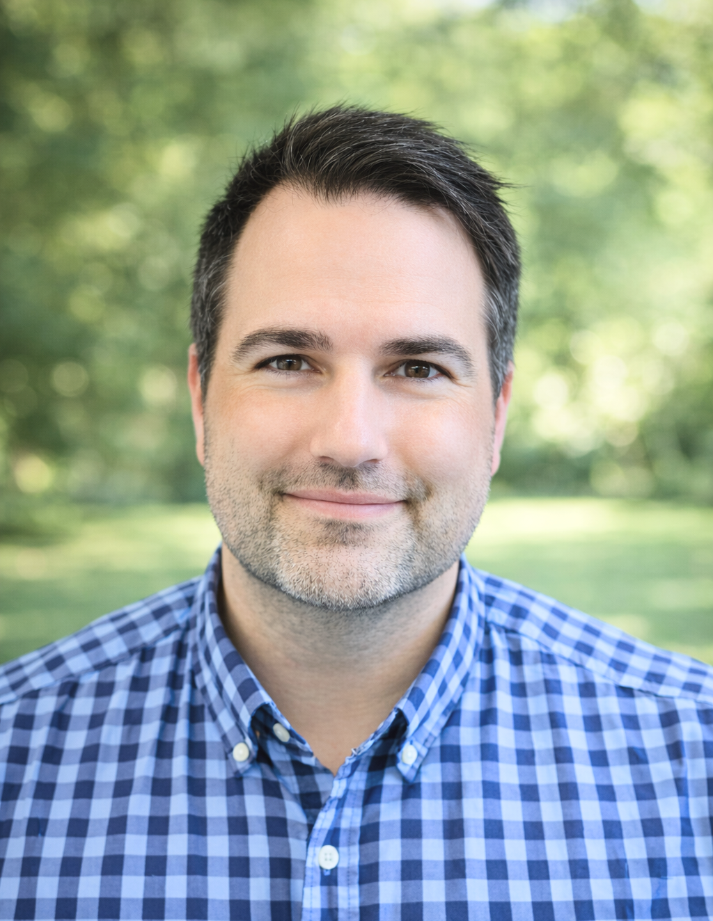 Headshot of a smiling man with dark hair and stubble, wearing a blue checkered shirt, outdoors with blurred green trees in the background.
