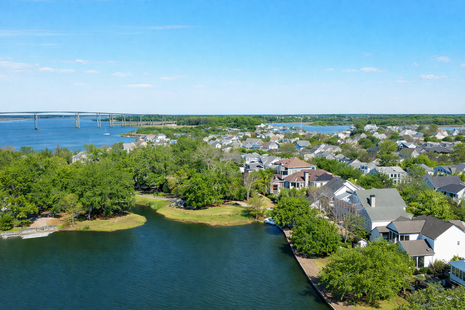 Aerial view of a lakeside neighborhood with houses, trees, and a water body, under a clear blue sky. A bridge is visible in the distance over the water.