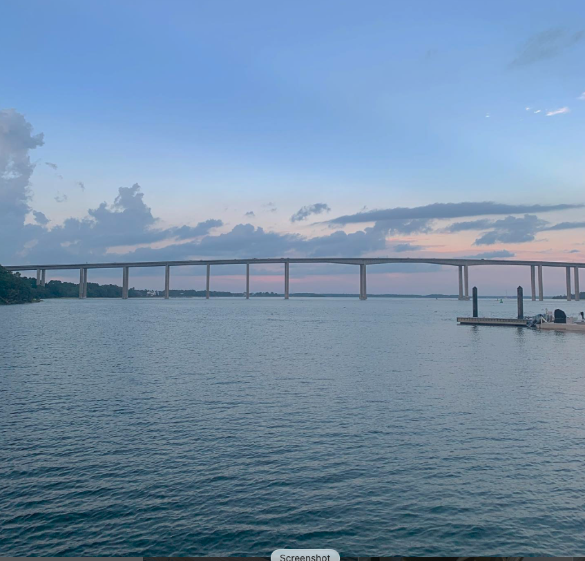 A large body of water with a bridge spanning across it and a small dock with boats on the right side. The sky has scattered clouds with a hint of sunset colors.
