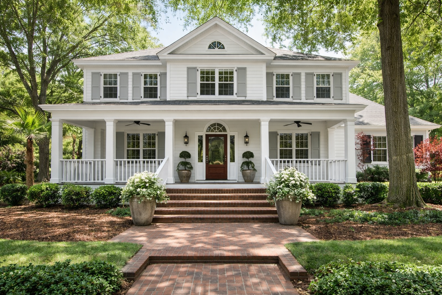 White two-story house with a porch, gray shutters, and a wooden front door, surrounded by greenery and trees.