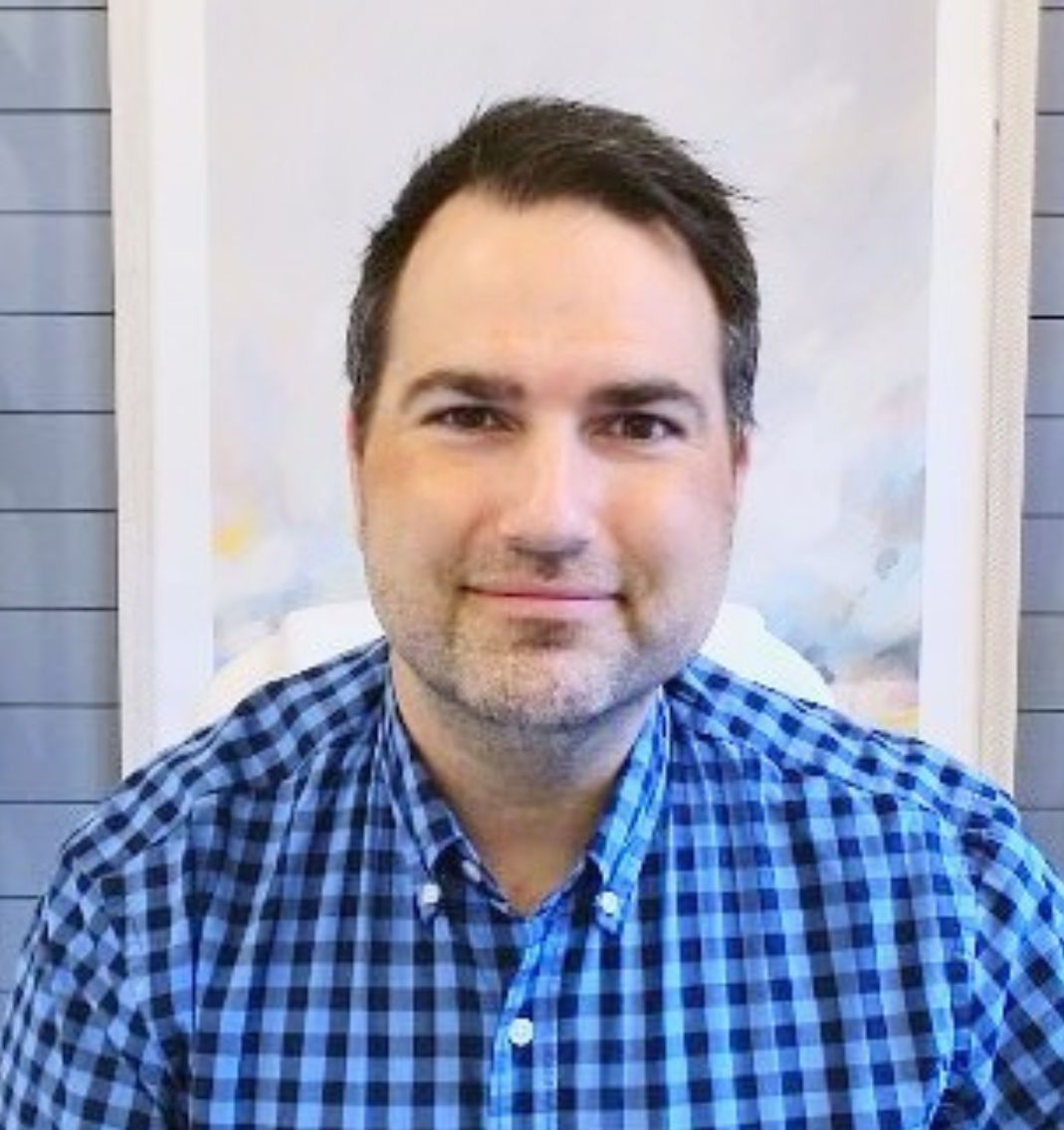 Portrait of a man with short dark hair, wearing a blue checkered shirt, smiling, with a blurred background.