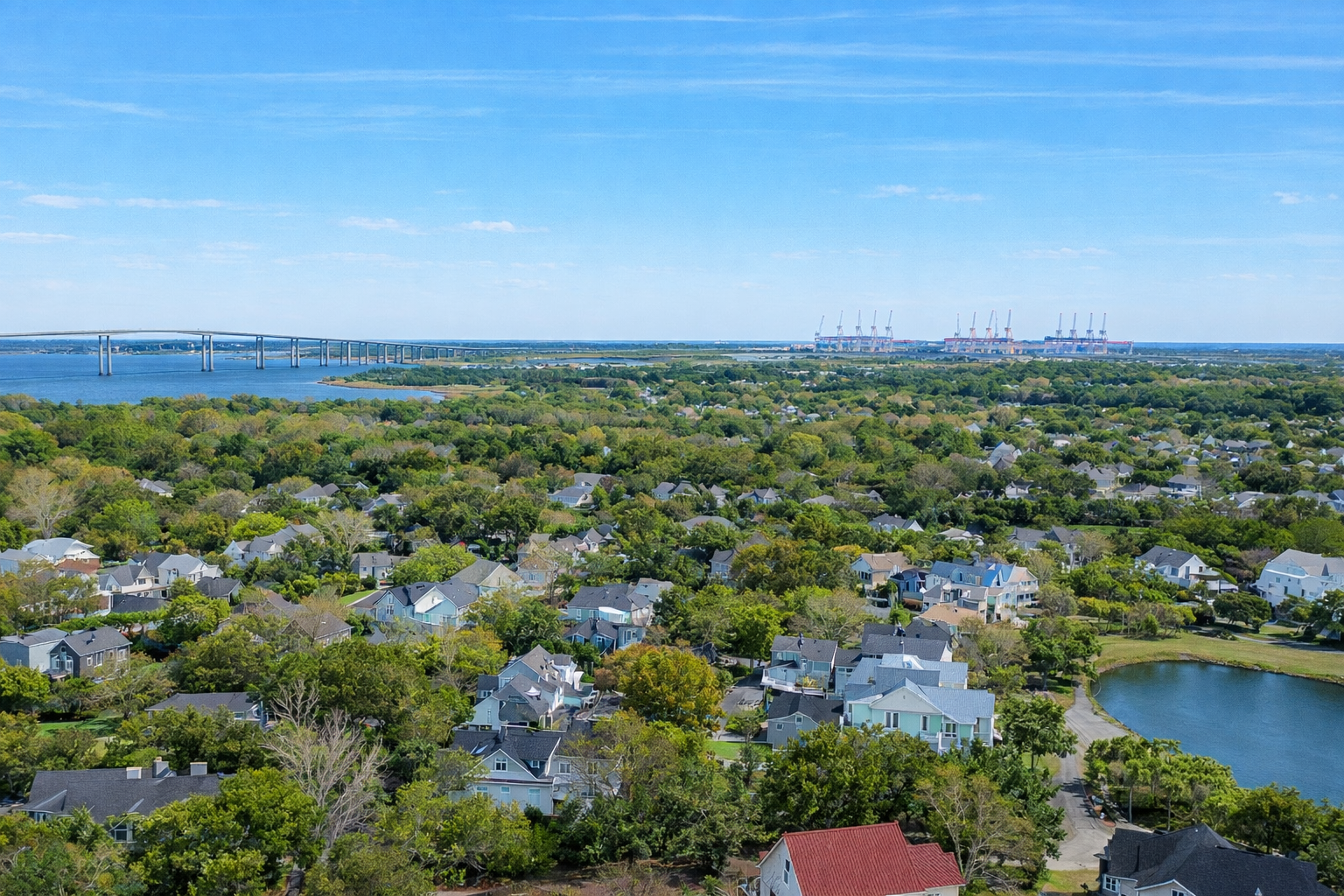 Aerial view of a suburban neighborhood near a body of water on a sunny day, with houses surrounded by green trees, a lake, a large bridge over the water, and industrial cranes in the distance under a blue sky.