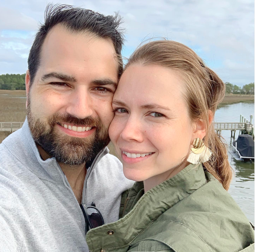 A smiling couple taking a close-up selfie outdoors near a body of water with a dock and boat in the background.
