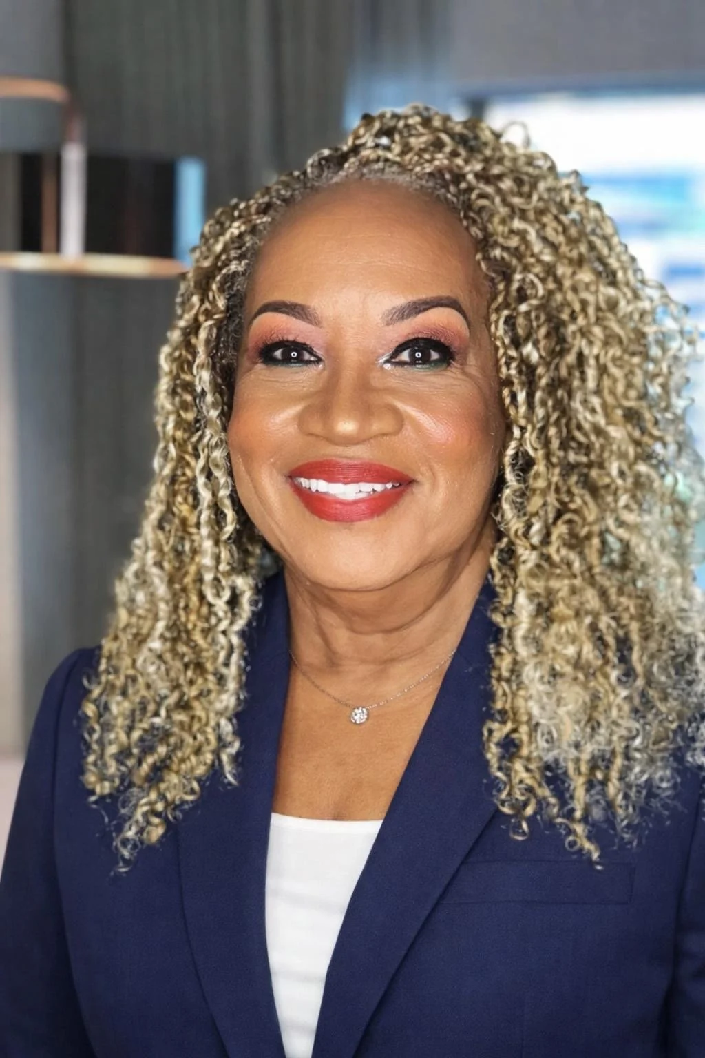 A woman with blonde curly hair smiling in an indoor setting, wearing a navy blazer and a necklace with a small pendant.