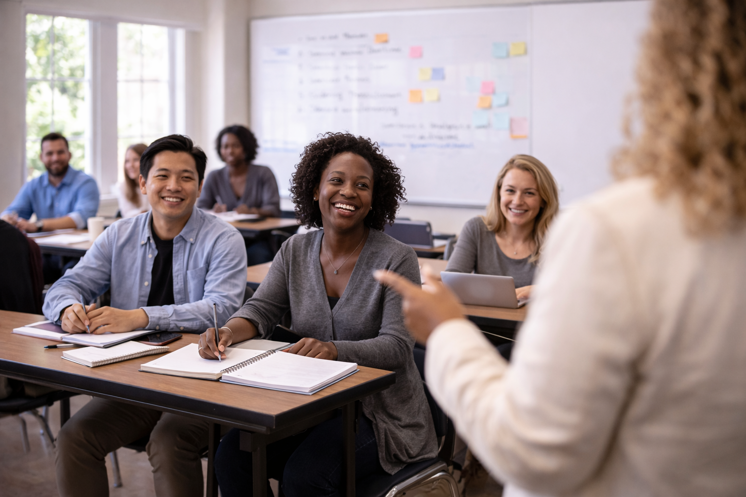 A diverse group of students in a classroom, smiling and listening to a woman instructor who is gesturing as she speaks.
