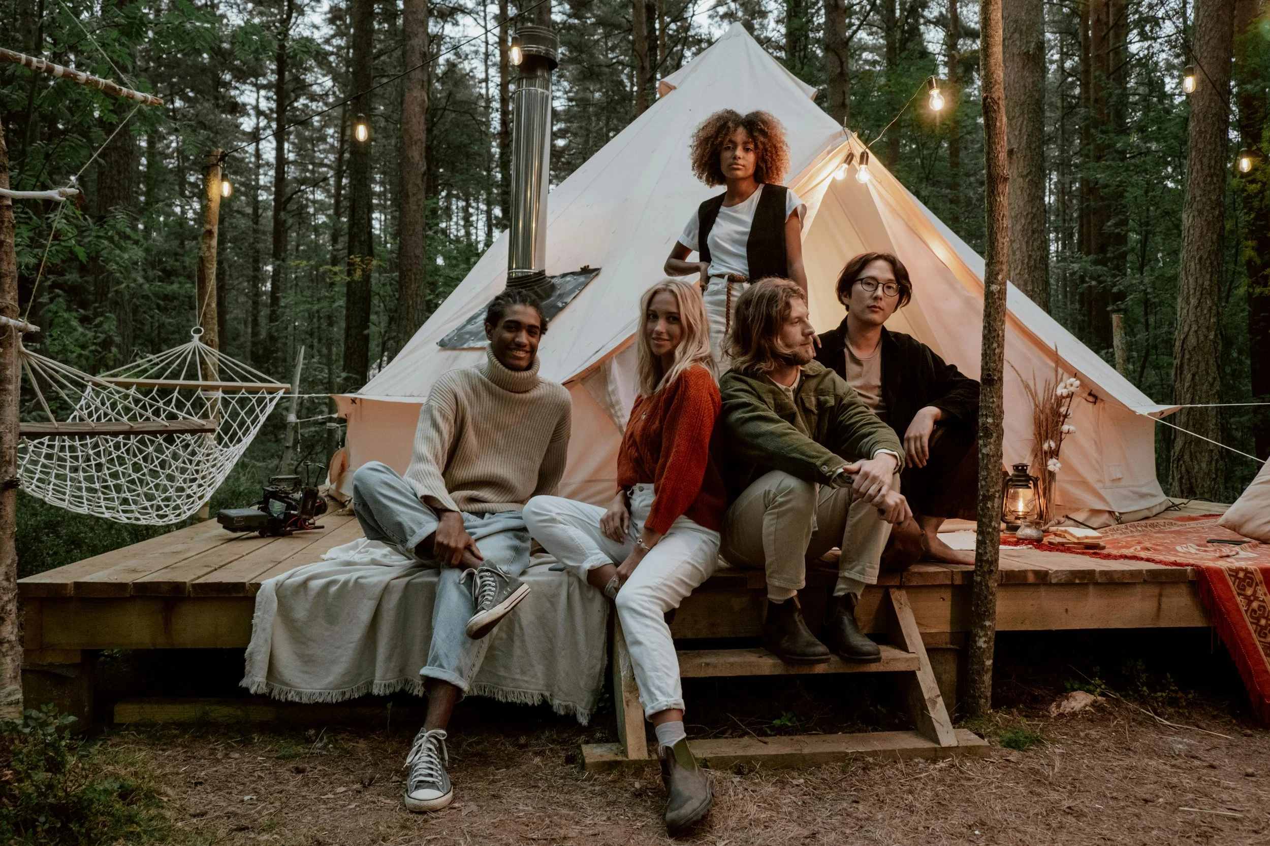 Group of five young adults sitting and standing in front of a large tent in a forest with string lights, hammock, and lanterns.