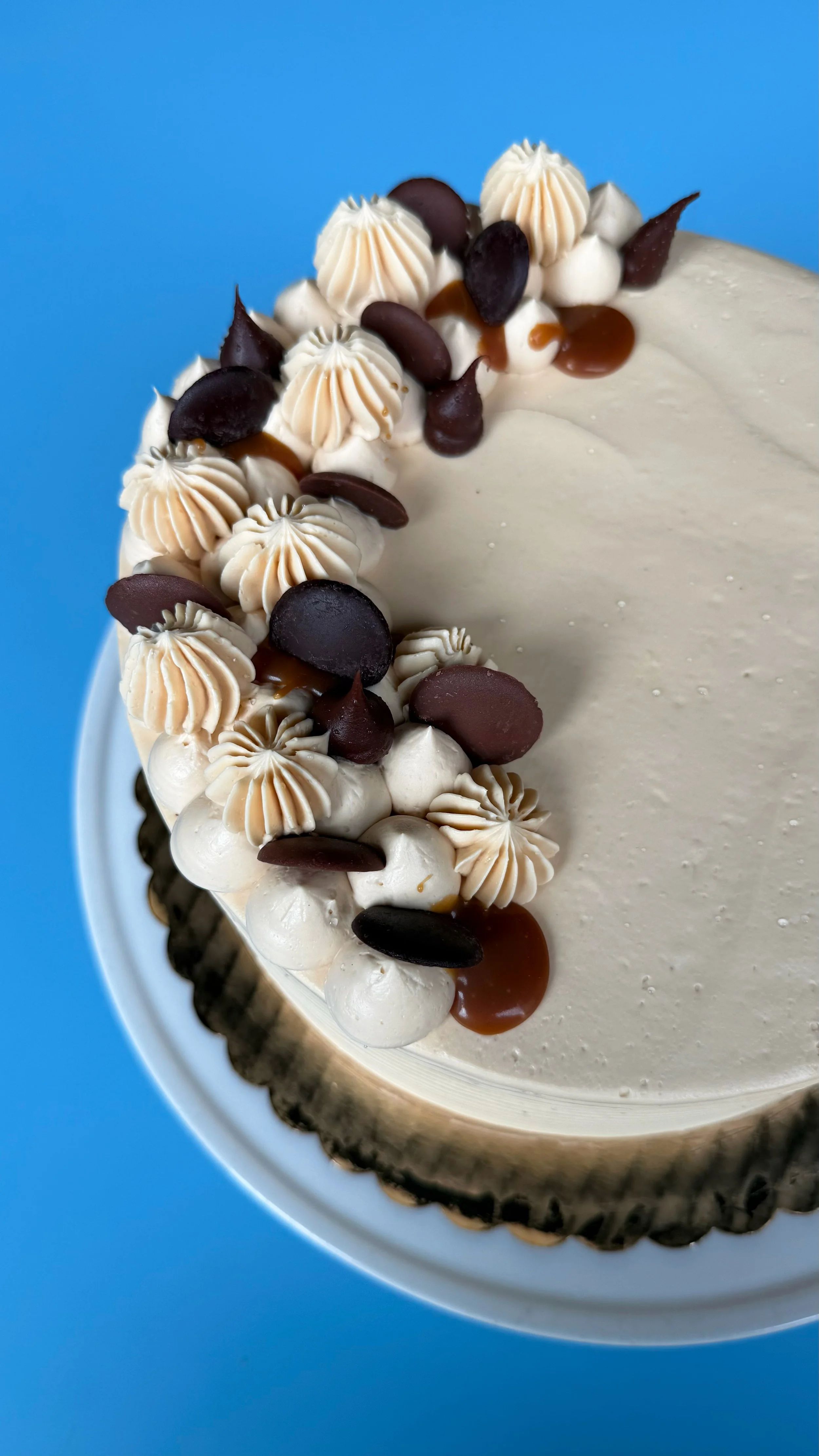 A close-up of a decorated birthday or celebration cake with white frosting, topped with dollops of cream, chocolate chips, and caramel drizzle on a blue background.