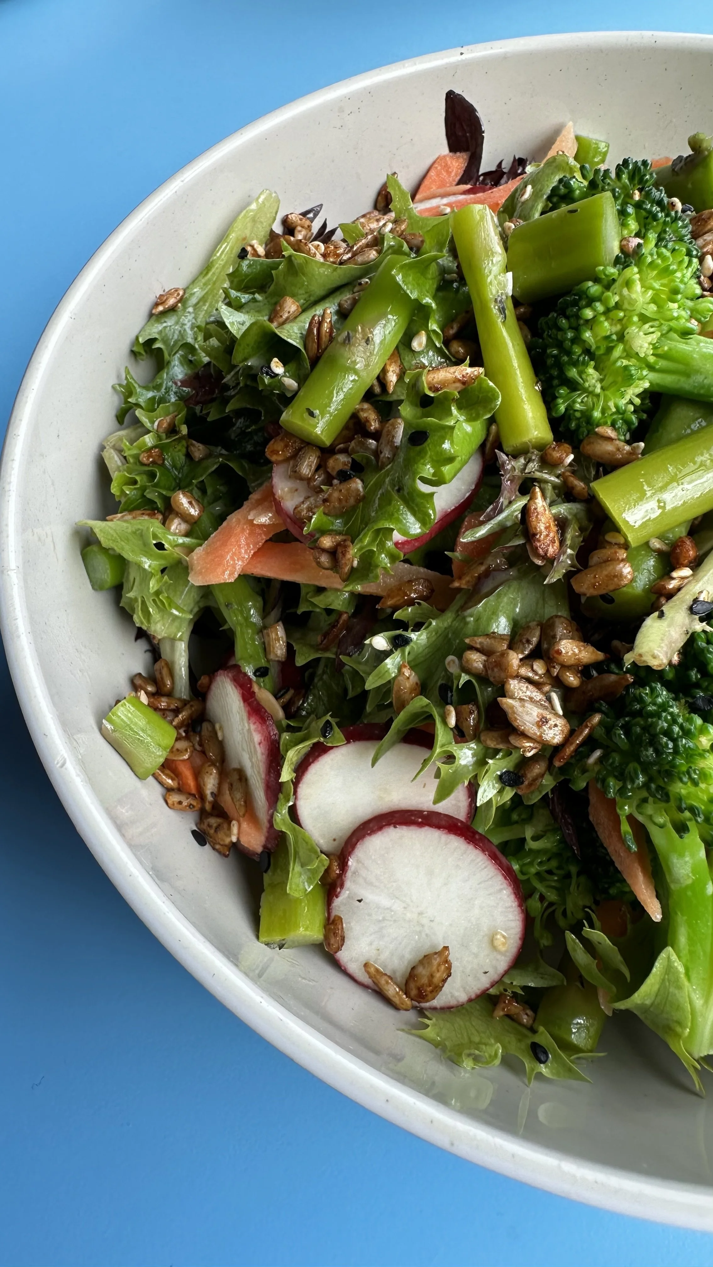 Fresh mixed green salad with radishes, broccoli, sunflower seeds, and chopped vegetables in a white bowl on a blue background.